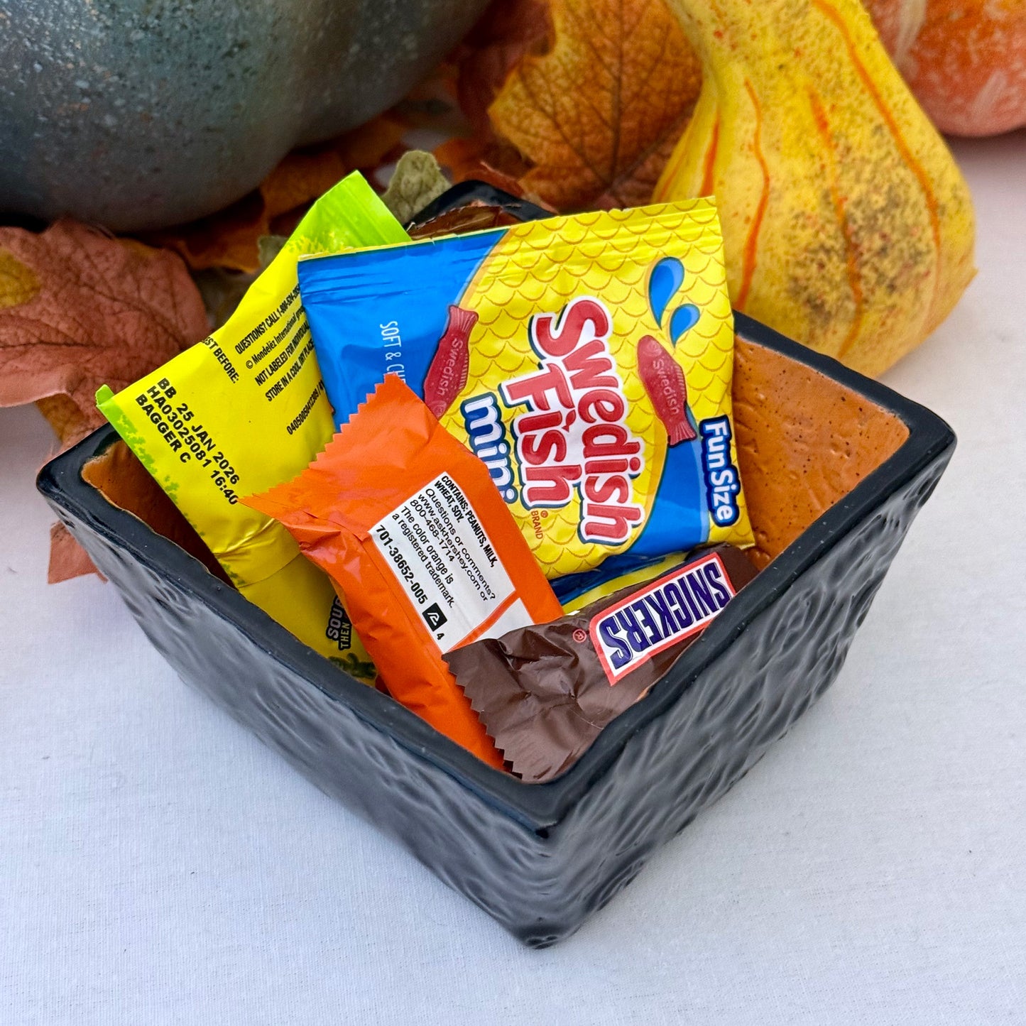 Square Halloween textured ceramic candy bowl with candy bars on a white surface with pumpkins in the background.