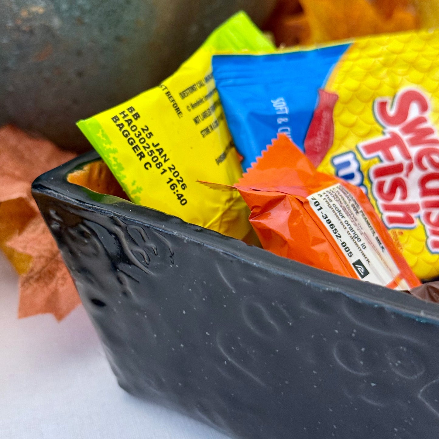 Square Halloween textured ceramic candy bowl with candy on a white surface with pumpkins in the background.