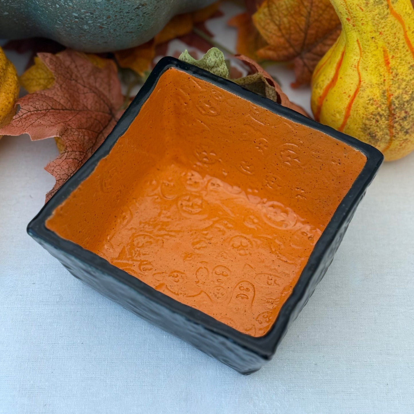 Square Halloween textured ceramic candy bowl on a white surface with pumpkins in the background.