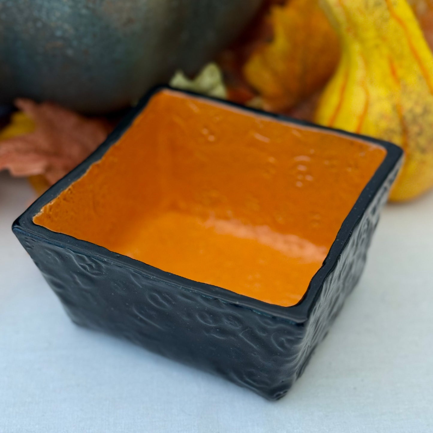 Square Halloween textured ceramic candy bowl on a white surface with pumpkins in the background.