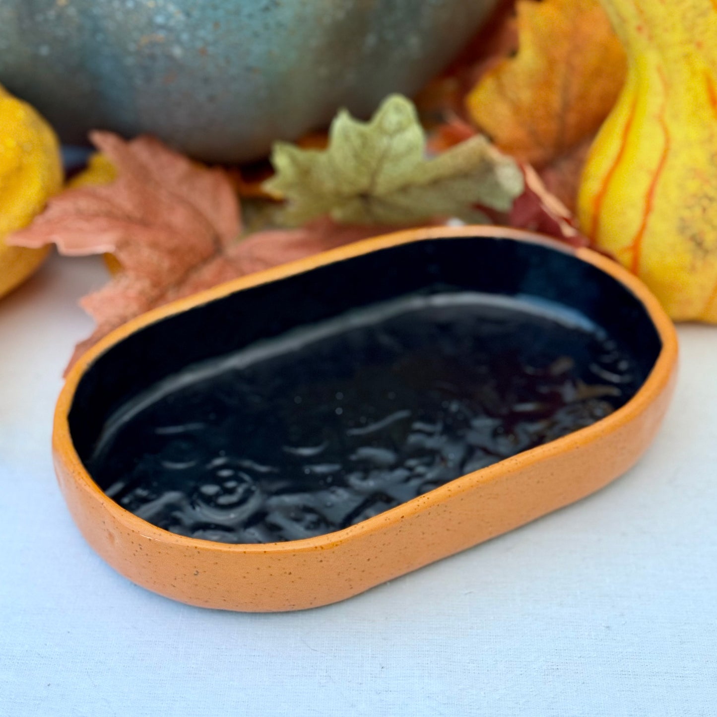 Black and orange Halloween textured oval trinket tray, surrounded by autumn leaves and a small pumpkin.