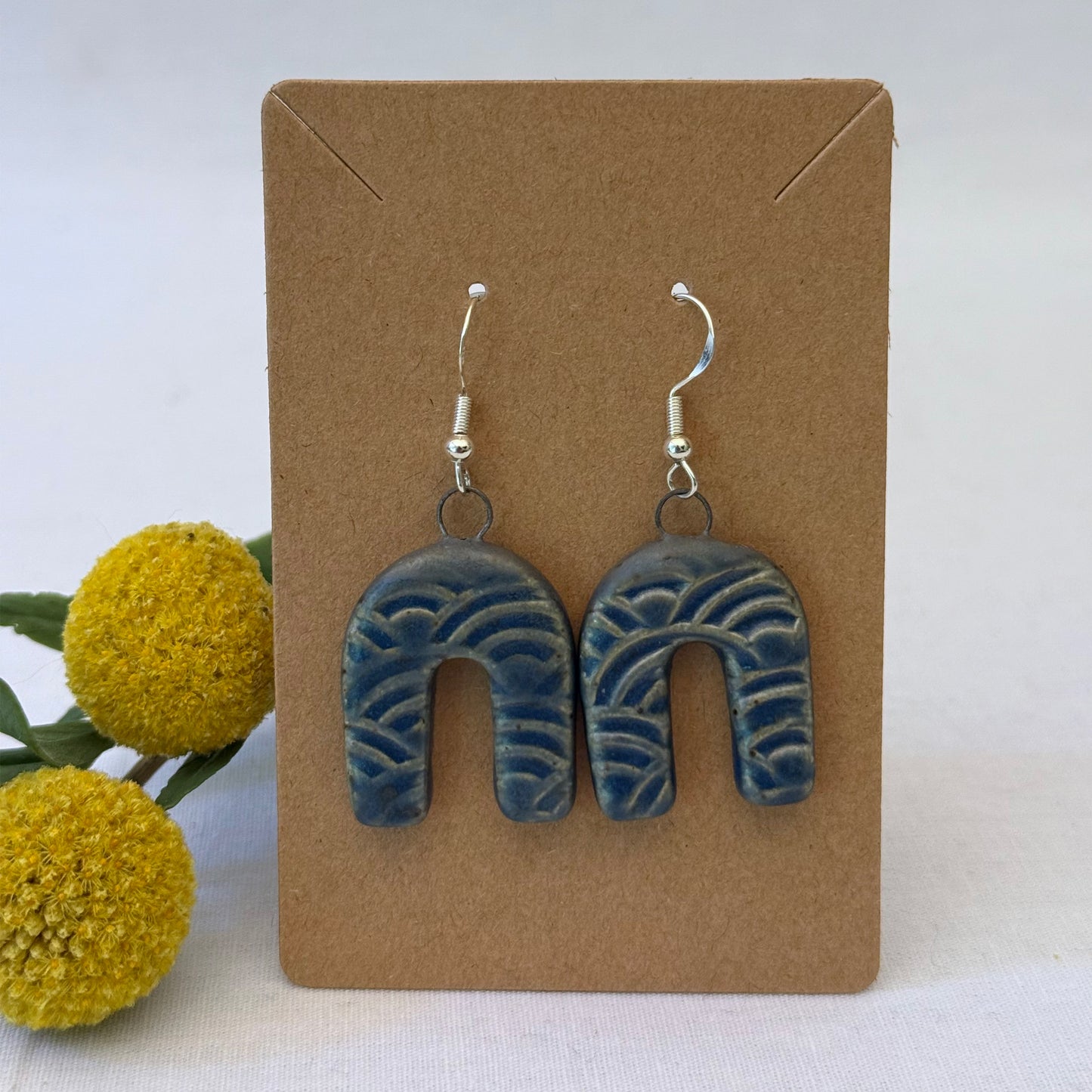 Blue wave-patterned earrings on a brown card with yellow flowers in the background