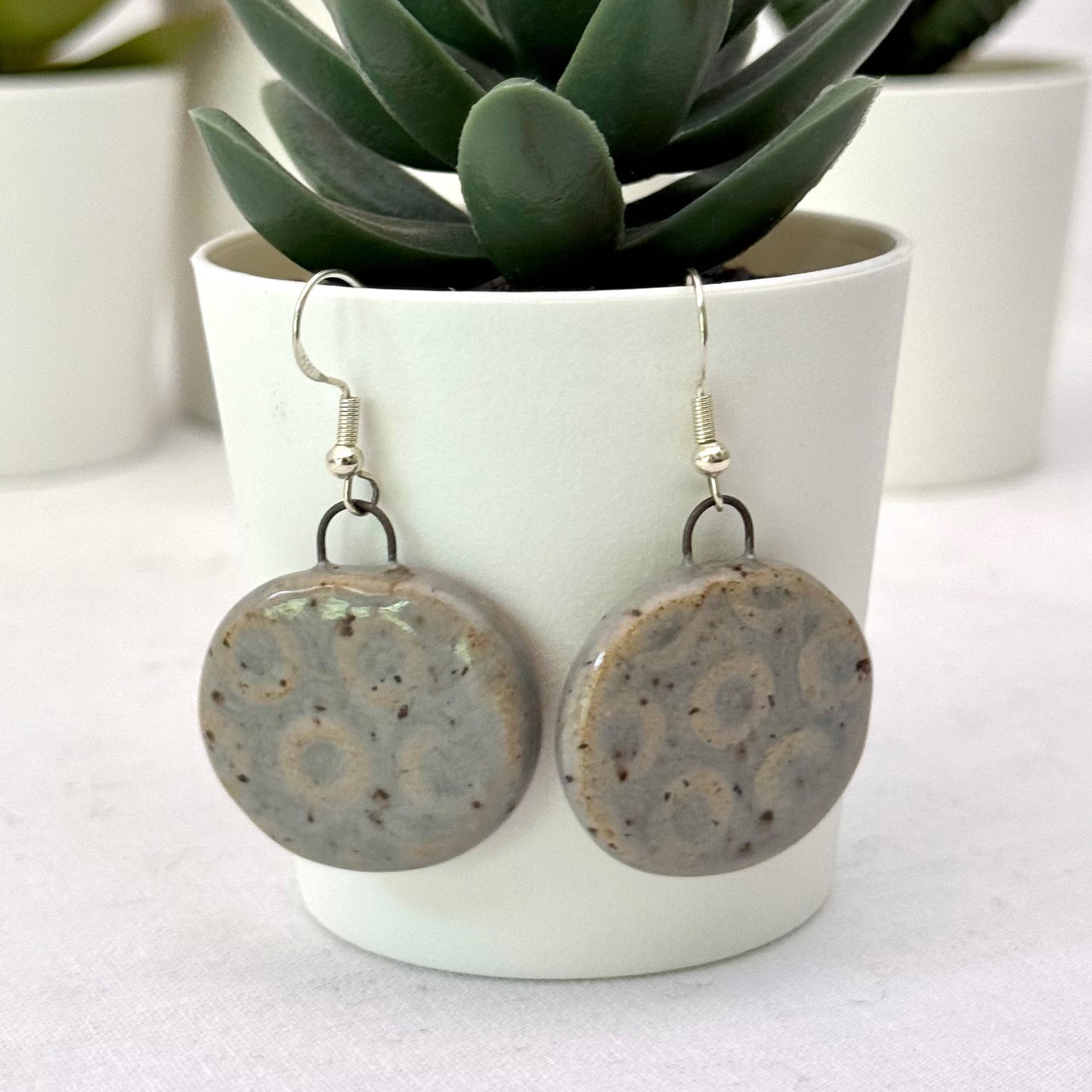 Pair of round grey textured earrings on a white surface with a plant in the background