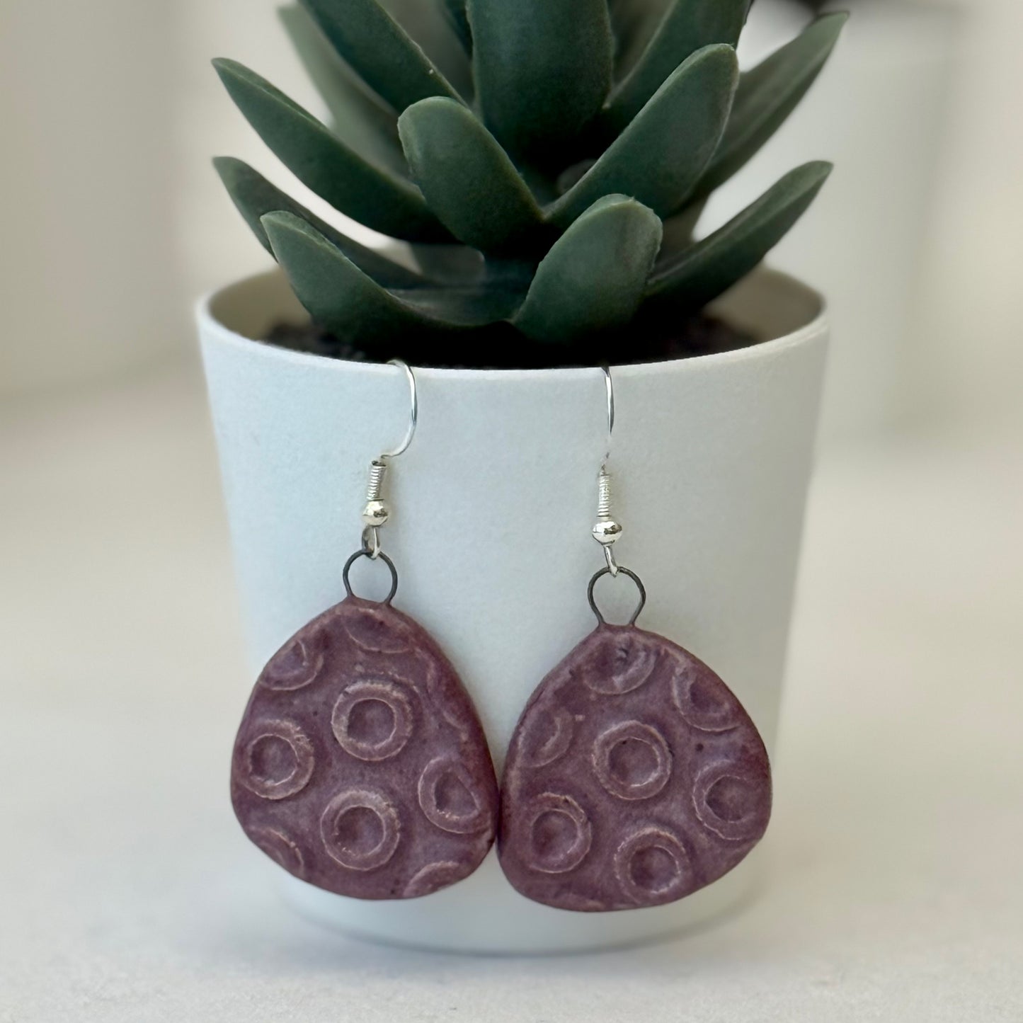 Purple textured earrings hanging in front of a white pot with a green plant.