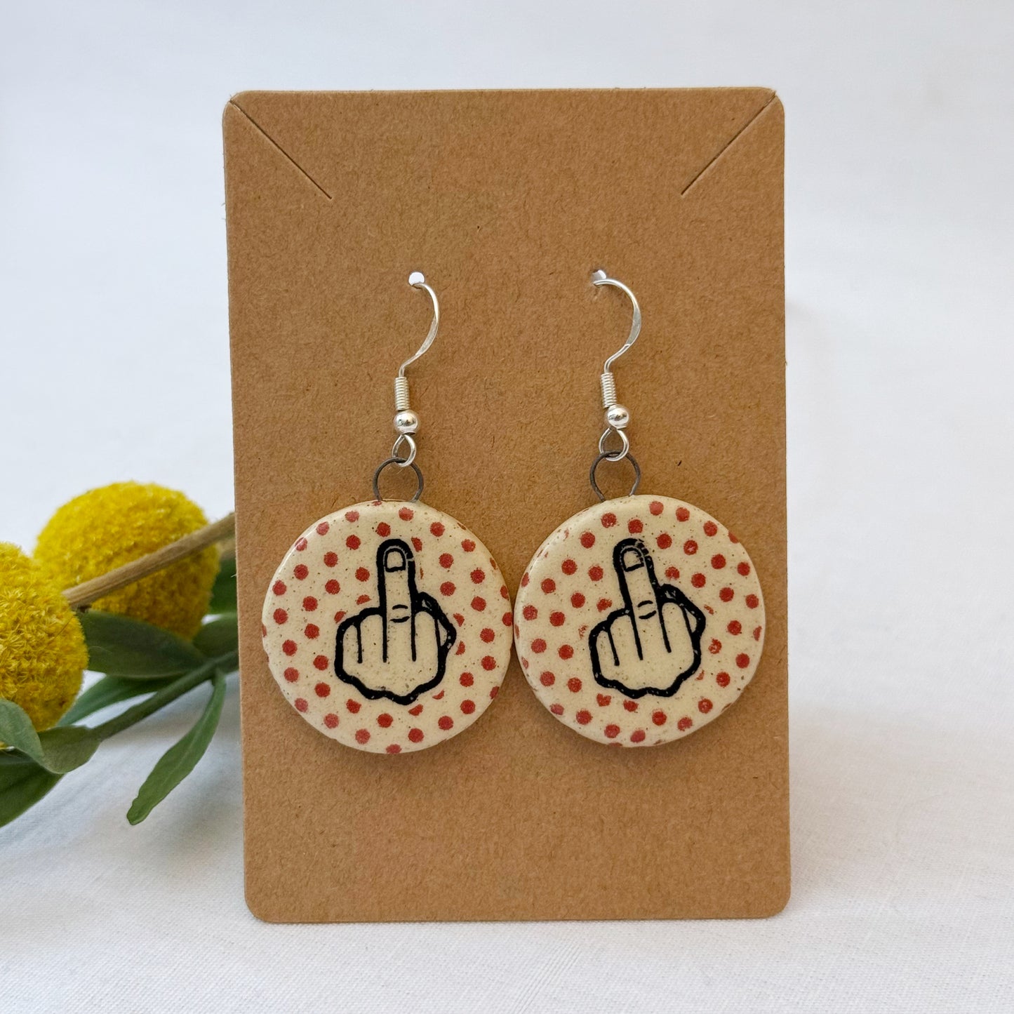 Earrings with middle finger design on polka dot background, displayed on a brown card.