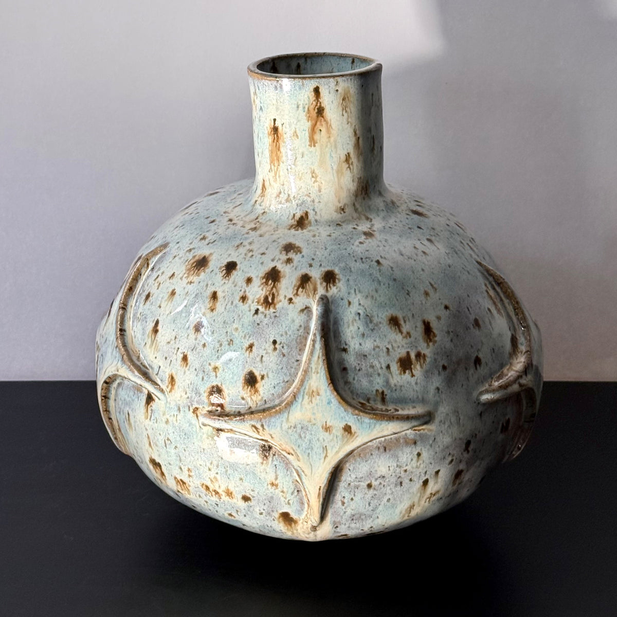 Handmade ceramic vase with a rounded sculptural body, narrow cylindrical neck, and raised atomic starburst motifs around the lower half. Finished in a pale blue-and-cream mottled glaze with darker speckled accents that highlight the carved geometric details.