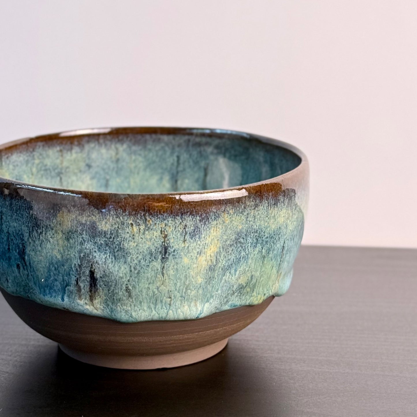 A handmade ceramic bowl with a textured, dripping glaze in shades of teal, blue, and brown, resting on a dark surface against a light background.