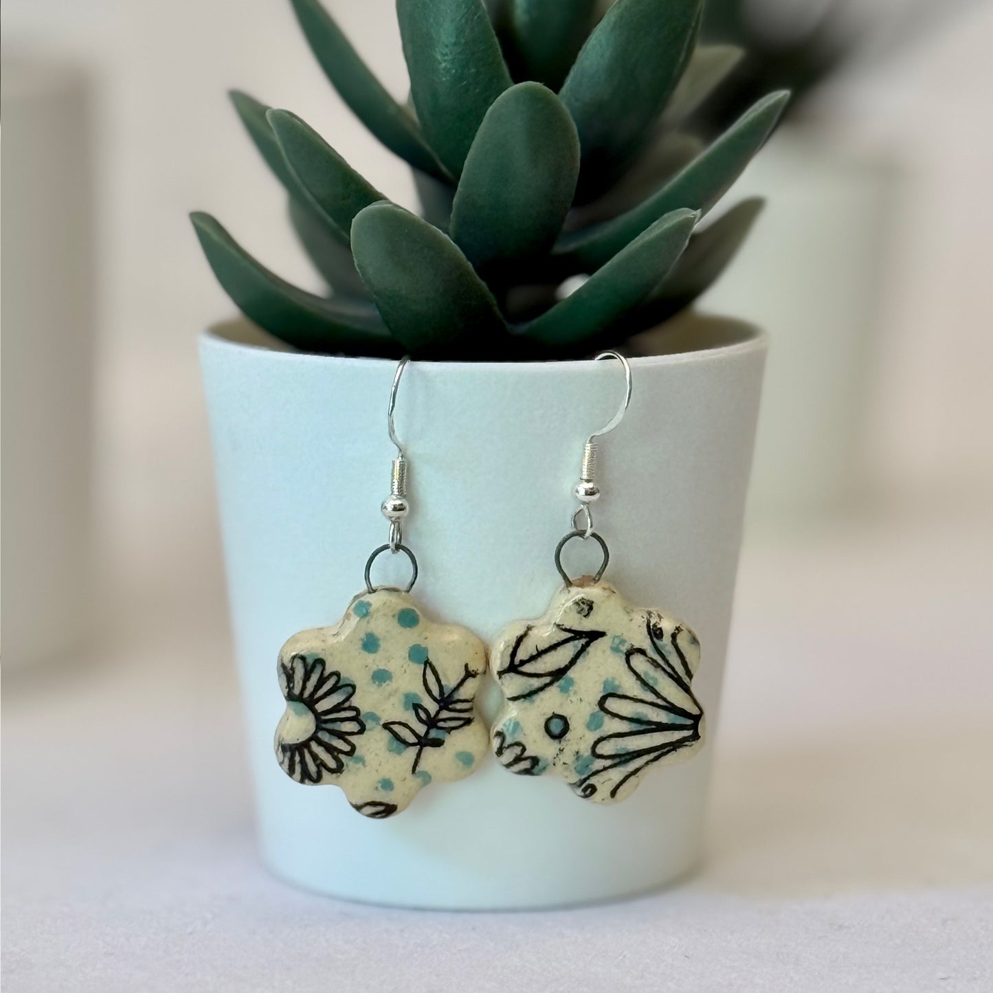 Pair of floral earrings on a white surface with a potted plant in the background