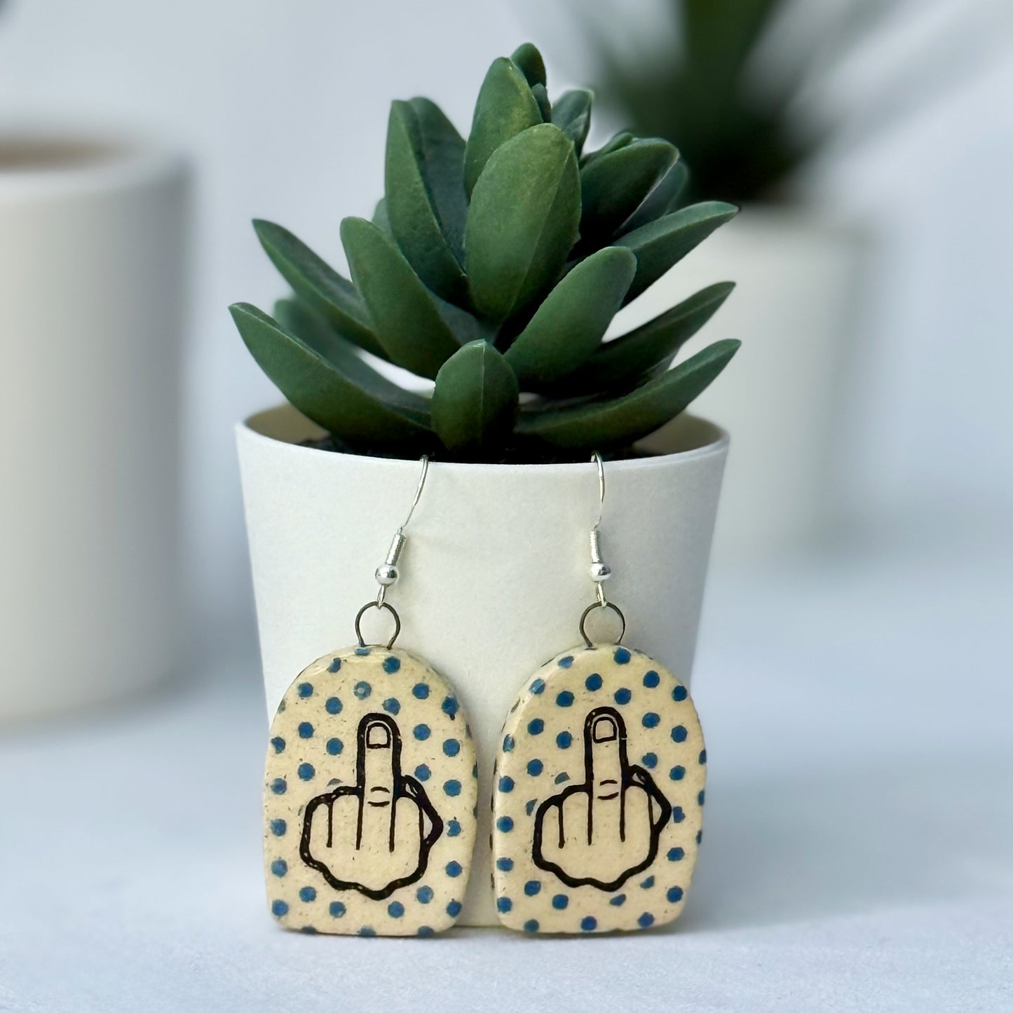 Earrings with middle finger design on polka dot background in front of a potted plant.