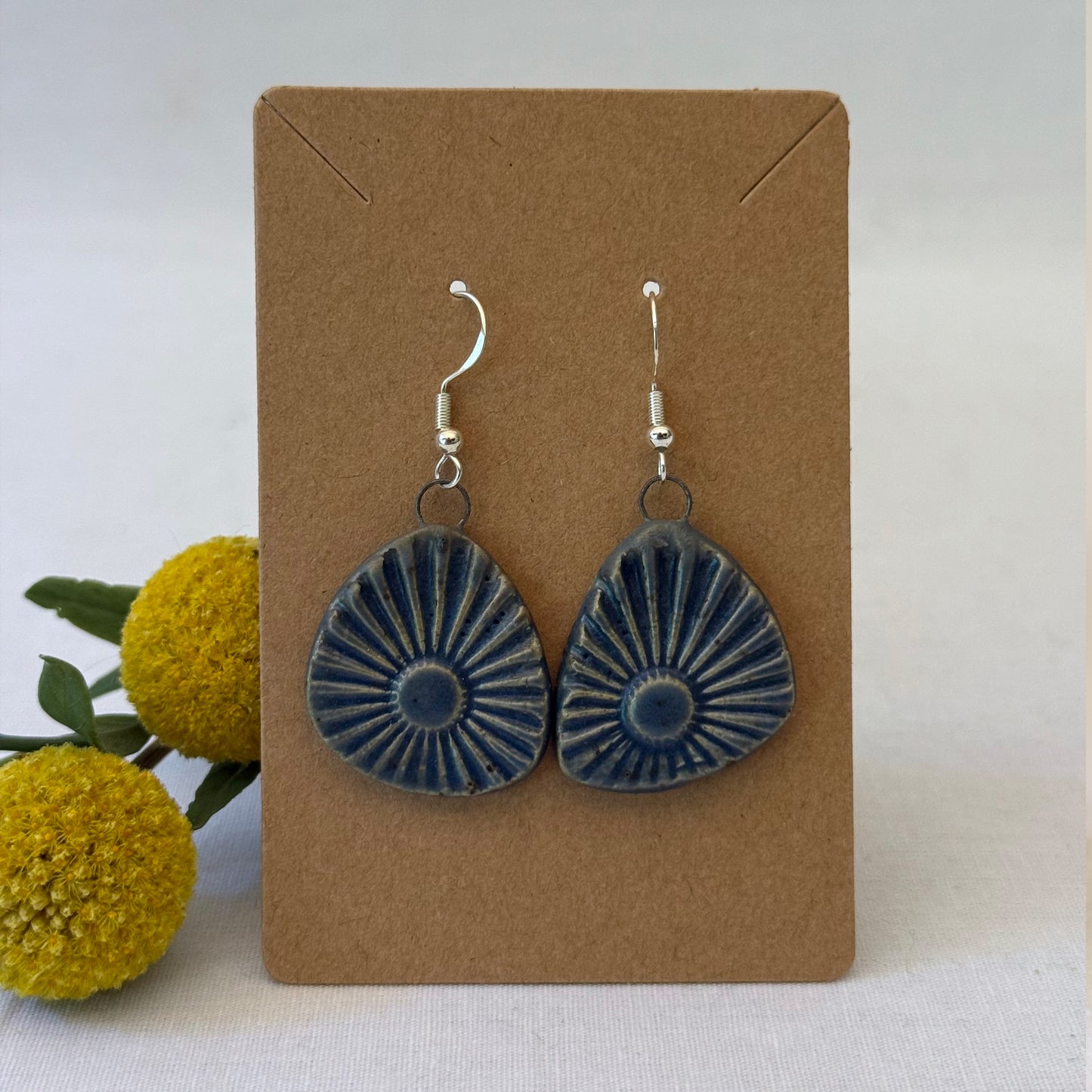 Blue ceramic earrings on a brown card with yellow flowers in the background