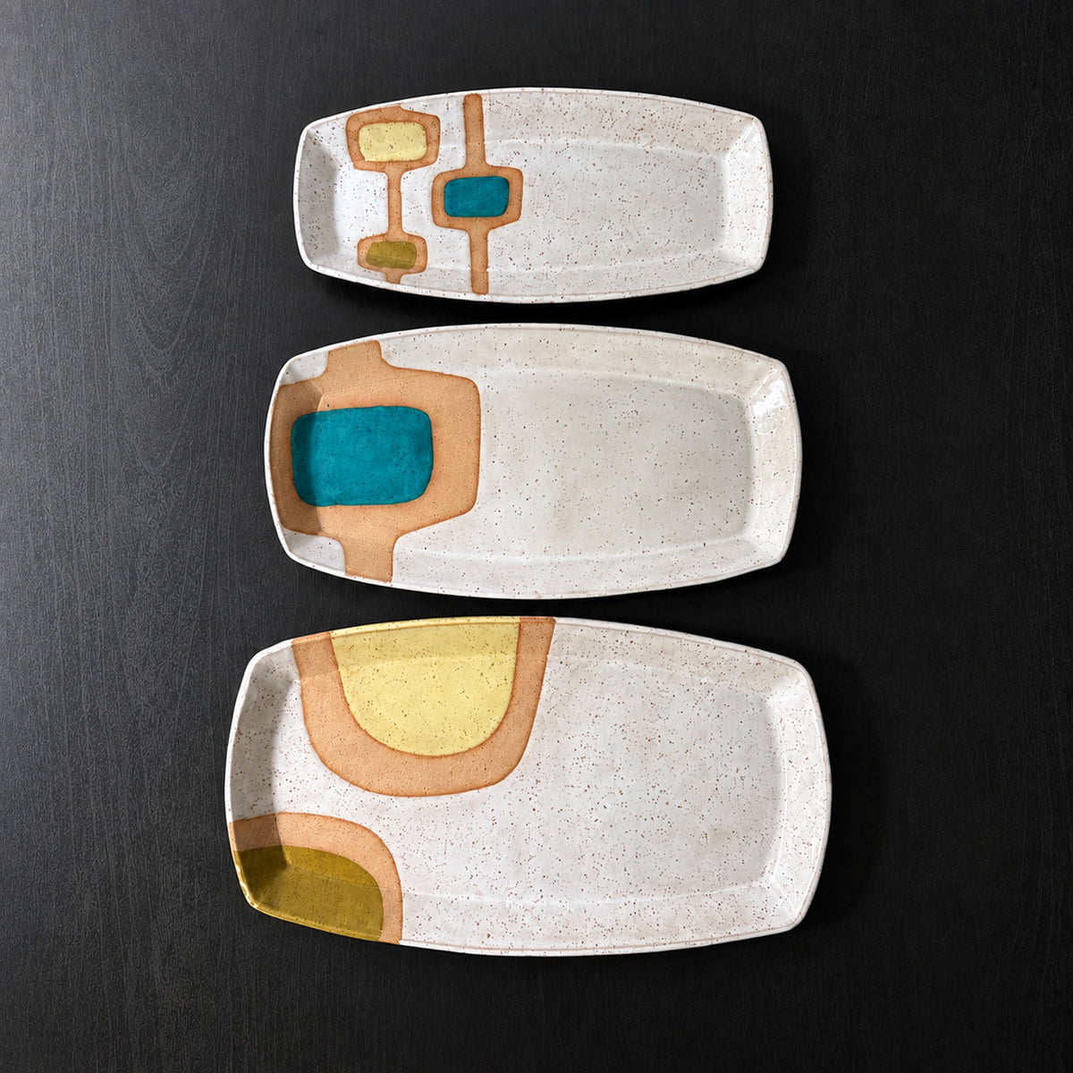 Three handmade rectangular ceramic trays with softly rounded corners, finished in a glossy white glaze with exposed speckled clay forming mid-century geometric designs. Accents of turquoise blue, mustard yellow, and olive green add pops of color across the three different patterns.