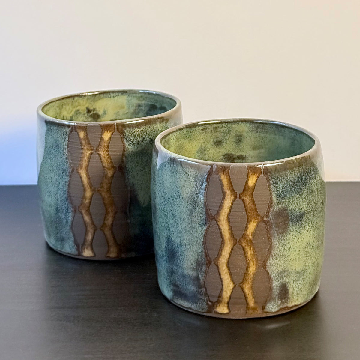 Handmade ceramic tumblers with a rounded shape and mottled glaze in mossy green, teal, blue, and warm ochre. A vertical geometric breeze-block motif on the front reveals areas of raw stoneware, creating a bold mid-century modern contrast.