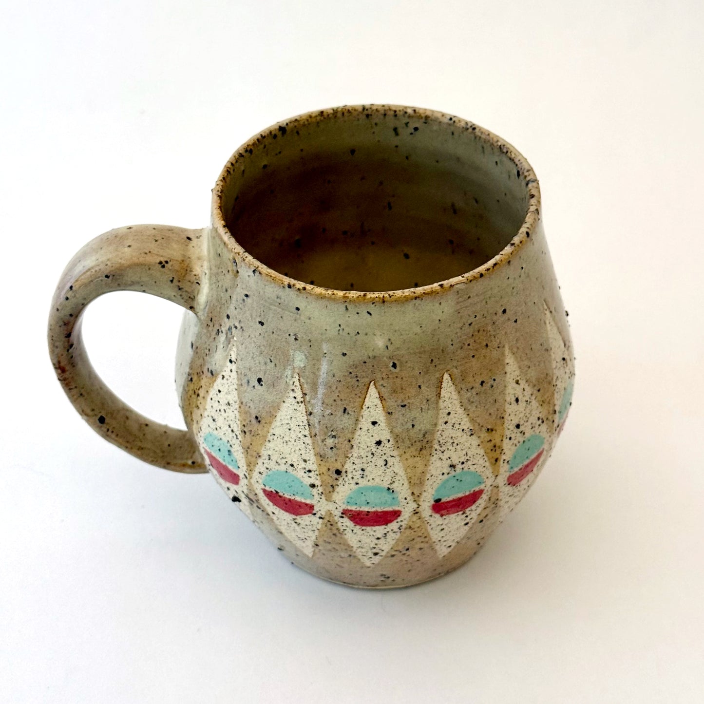 A bell-shaped, speckled tan stoneware mug featuring a pattern of cream diamonds. Each diamond contains a circular motif split into teal and poppy red. The mug has a large, rounded handle and visible dark mineral flecks.