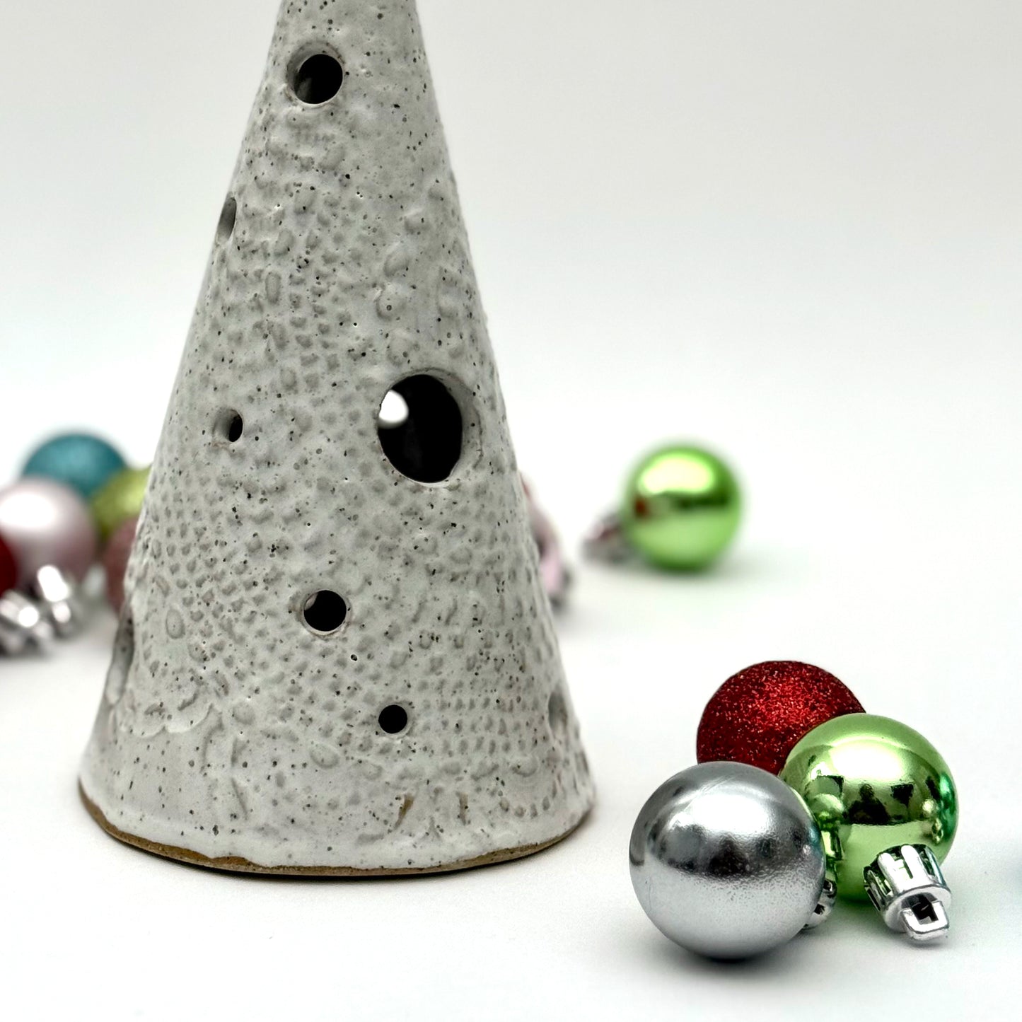 A white speckled ceramic tree-shaped luminary with light openings, styled among colorful holiday ornaments.