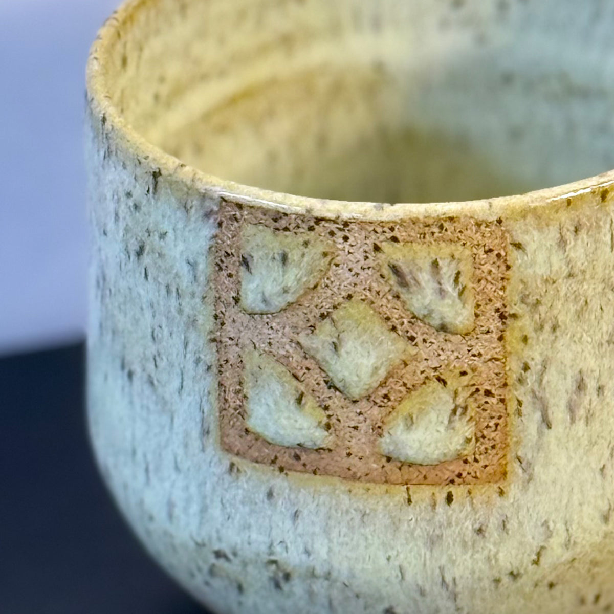 Close up of a handmade ceramic tumbler with a rounded shape and warm, yellow glaze. A square Empress breeze-block motif is inset on the front, revealing the natural clay and adding a mid-century geometric detail.
