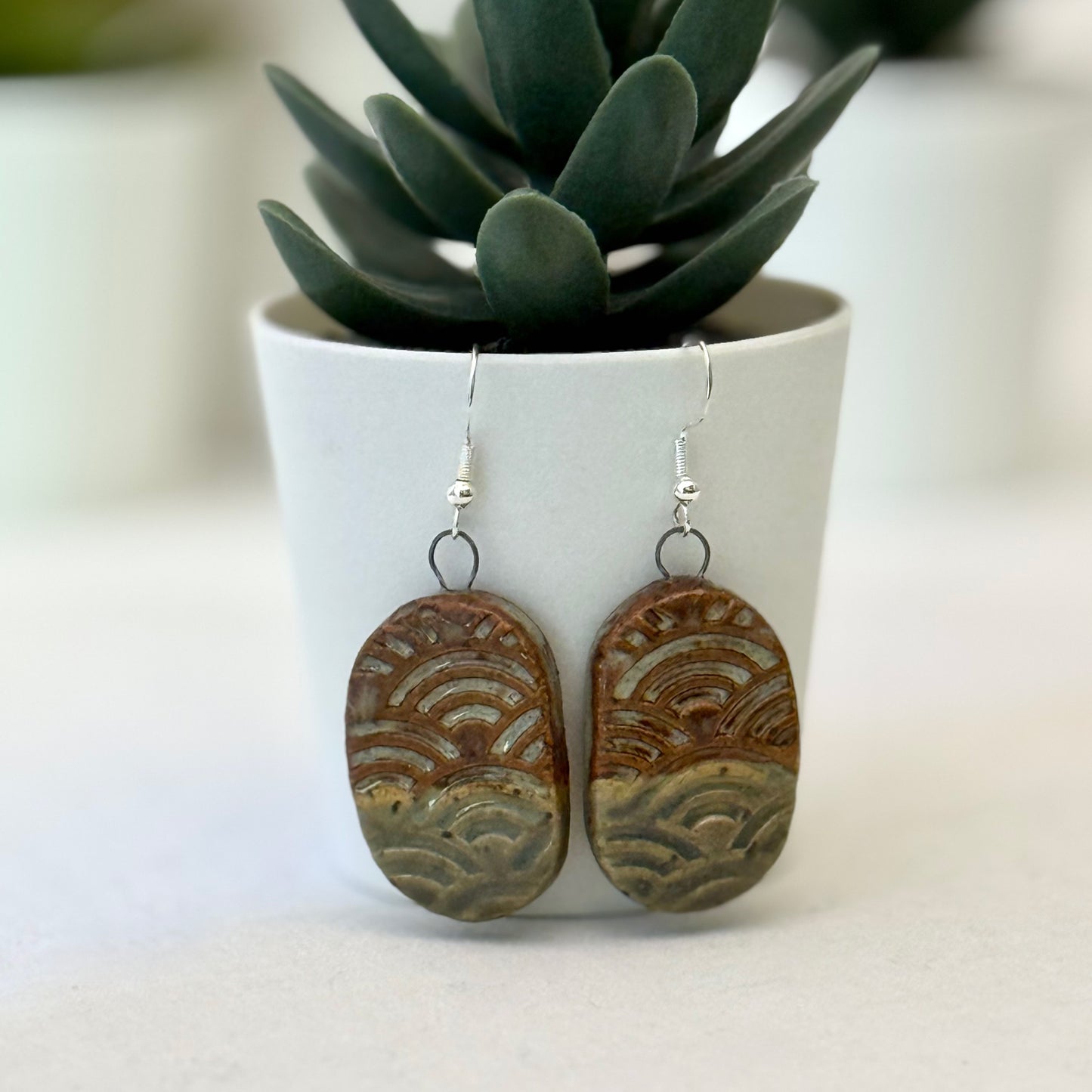 Pair of ceramic earrings with wave patterns in front of a potted plant.