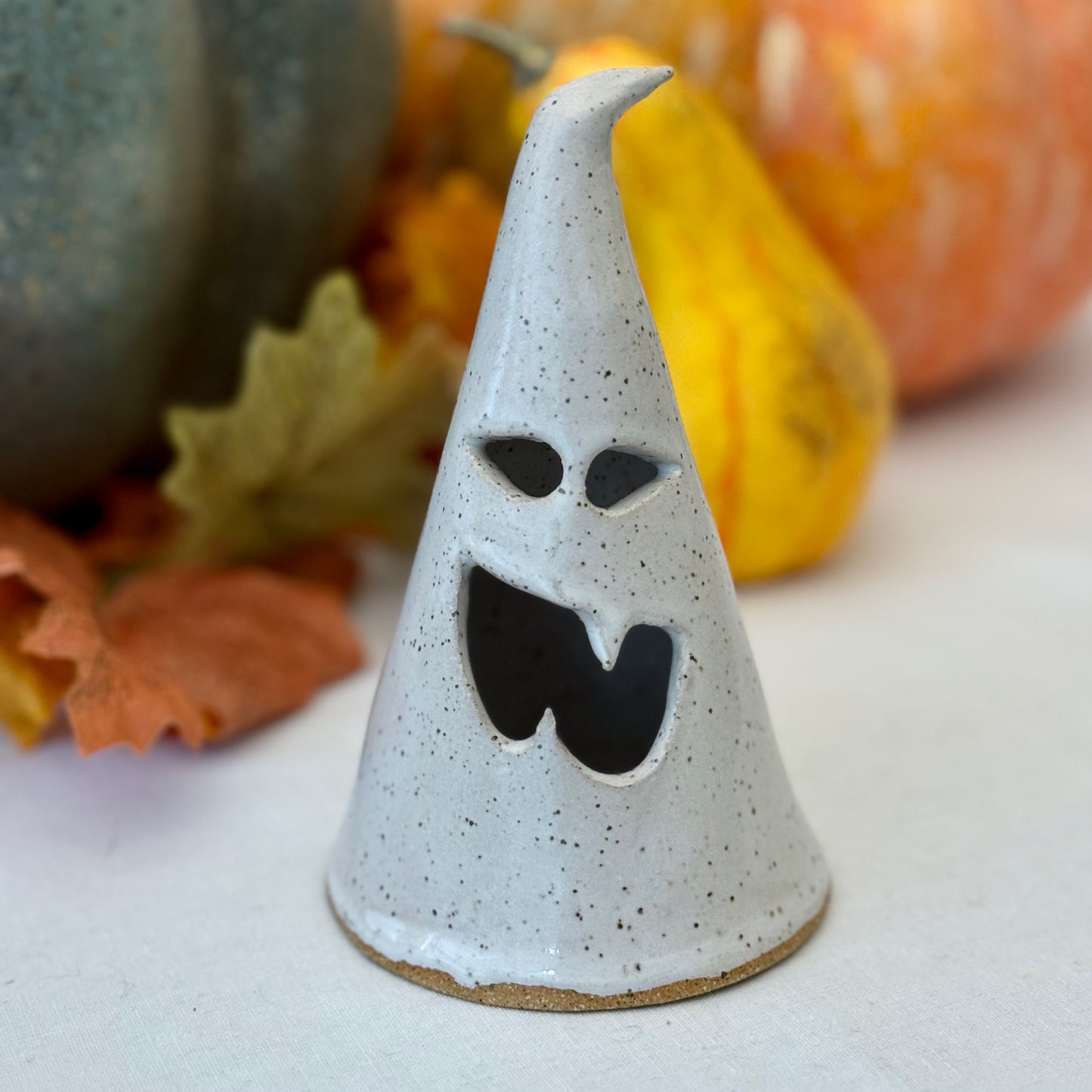 small ceramic ghost luminary on a white surface with pumpkins in the background.