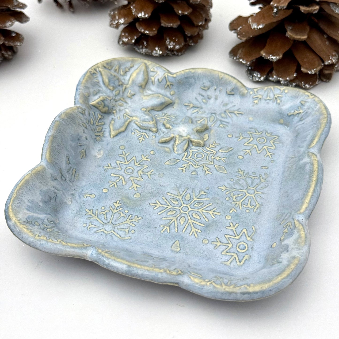 A handmade blue ceramic dish with scalloped edges and raised snowflake patterns where the glaze breaks to reveal white clay, shown with decorative pinecones.