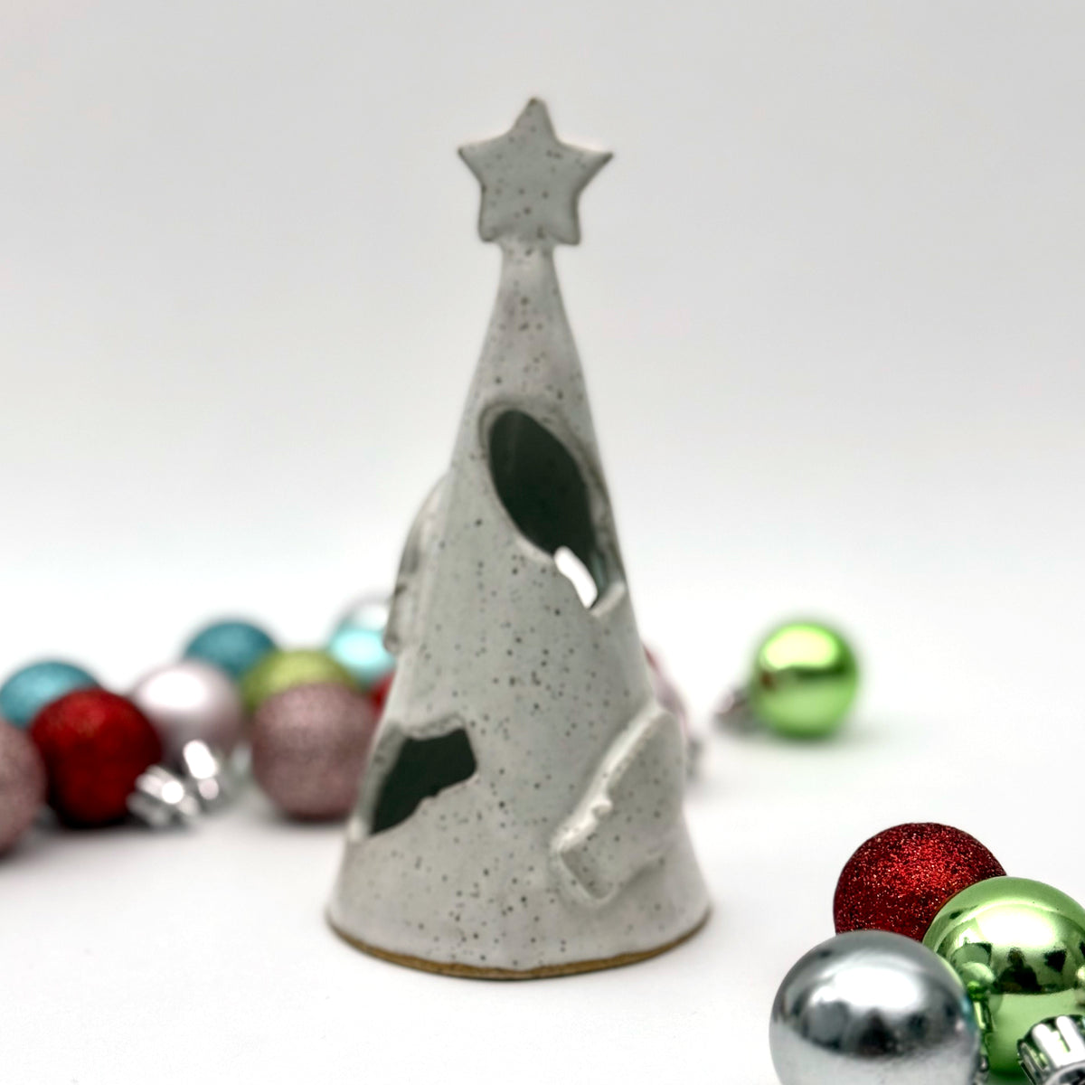 A white speckled ceramic tree luminary with cutout holiday light shapes and a star on top, surrounded by small colorful ornaments.