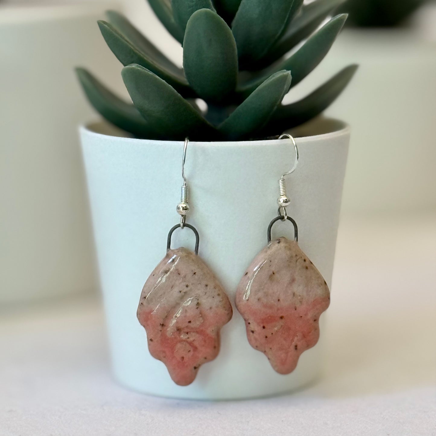 Pink ceramic earrings in front of a white pot with a plant.
