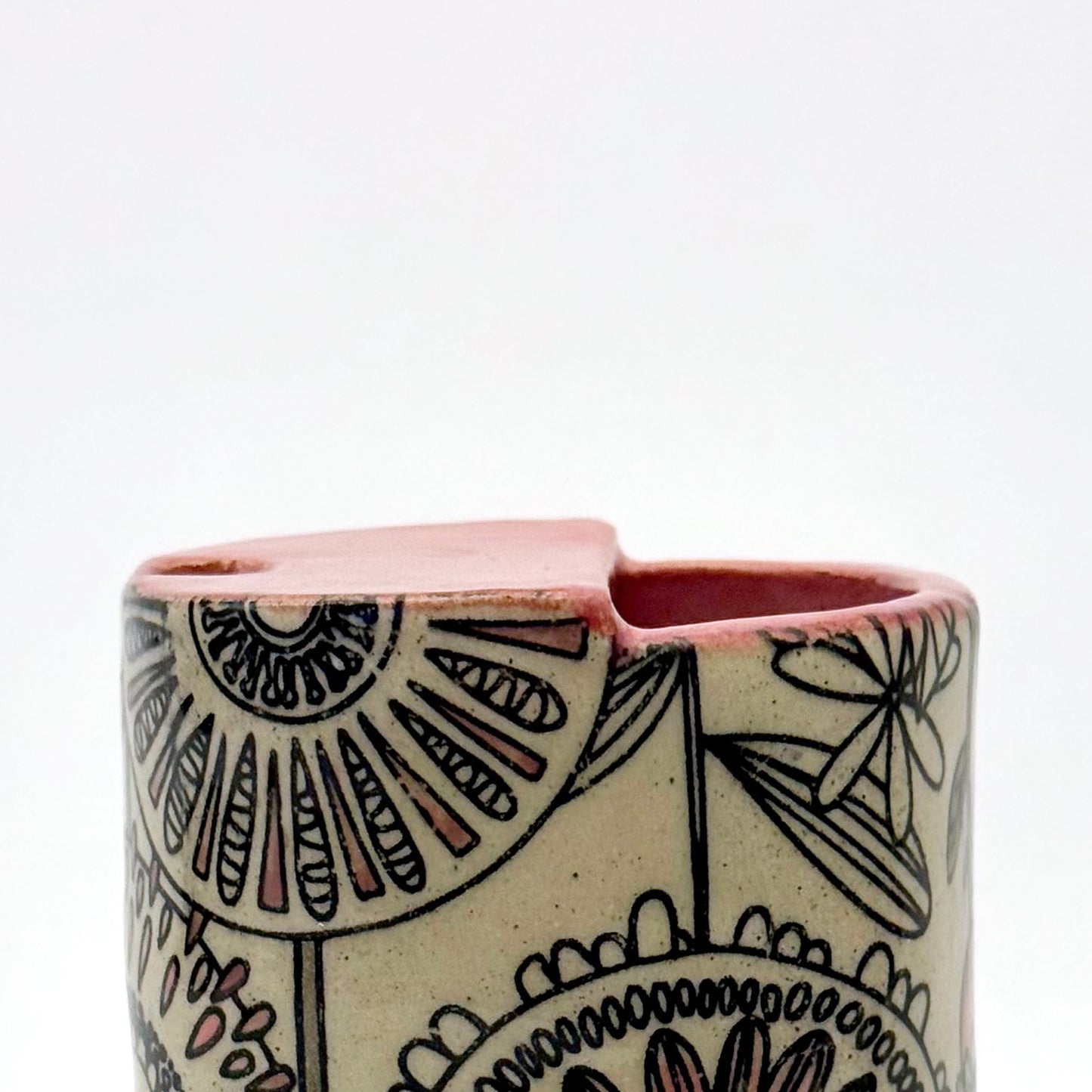 Close-up of handmade 14oz stoneware mug with mid-century inspired floral pattern and warm coral pink glazed top. The graphic flower design wraps around the exterior, and the mug features a cutout sip opening.