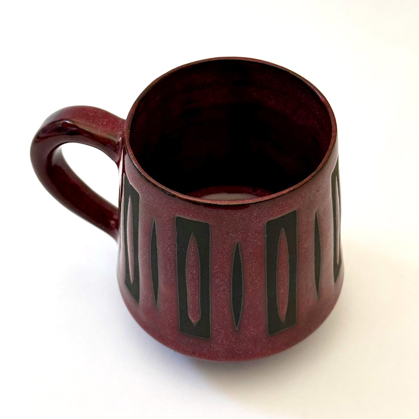Hand-thrown stoneware mug with a deep burgundy glaze and repeating elongated geometric pattern in dark teal-black. The bold mid-century-inspired design wraps around the slightly tapered mug, which features a curved handle and glossy finish. Photographed against a soft white background.