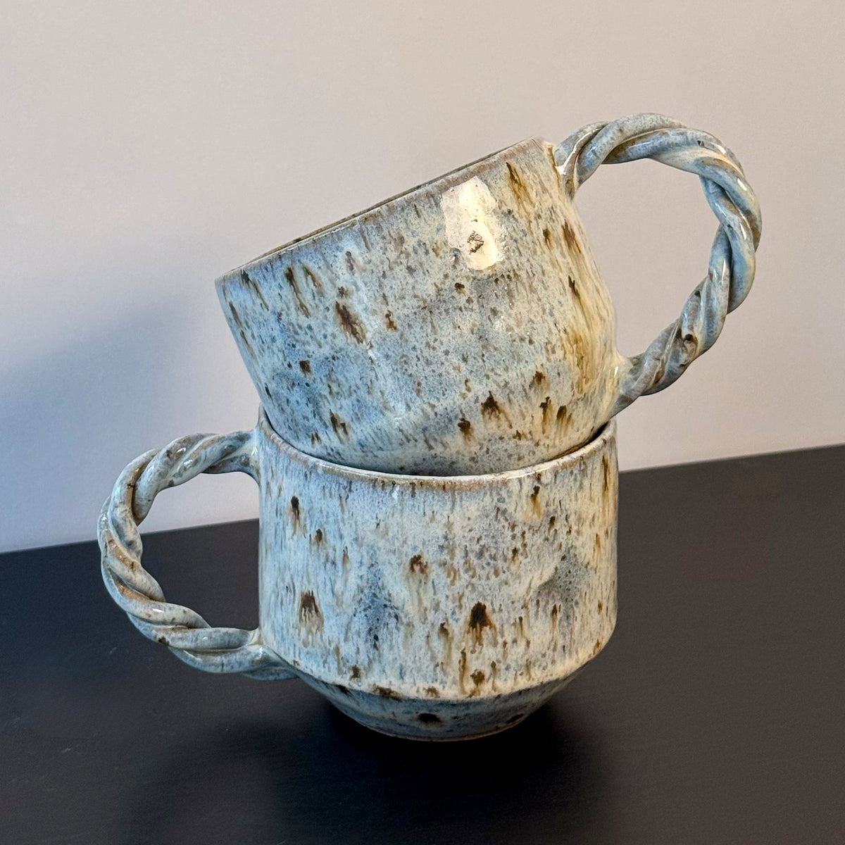 Two handmade ceramic mugs with rounded forms, soft mottled blue glaze, and chunky twisted handles. Each mug has a speckled stoneware surface and an organic, sculptural shape with subtle mid-century character.