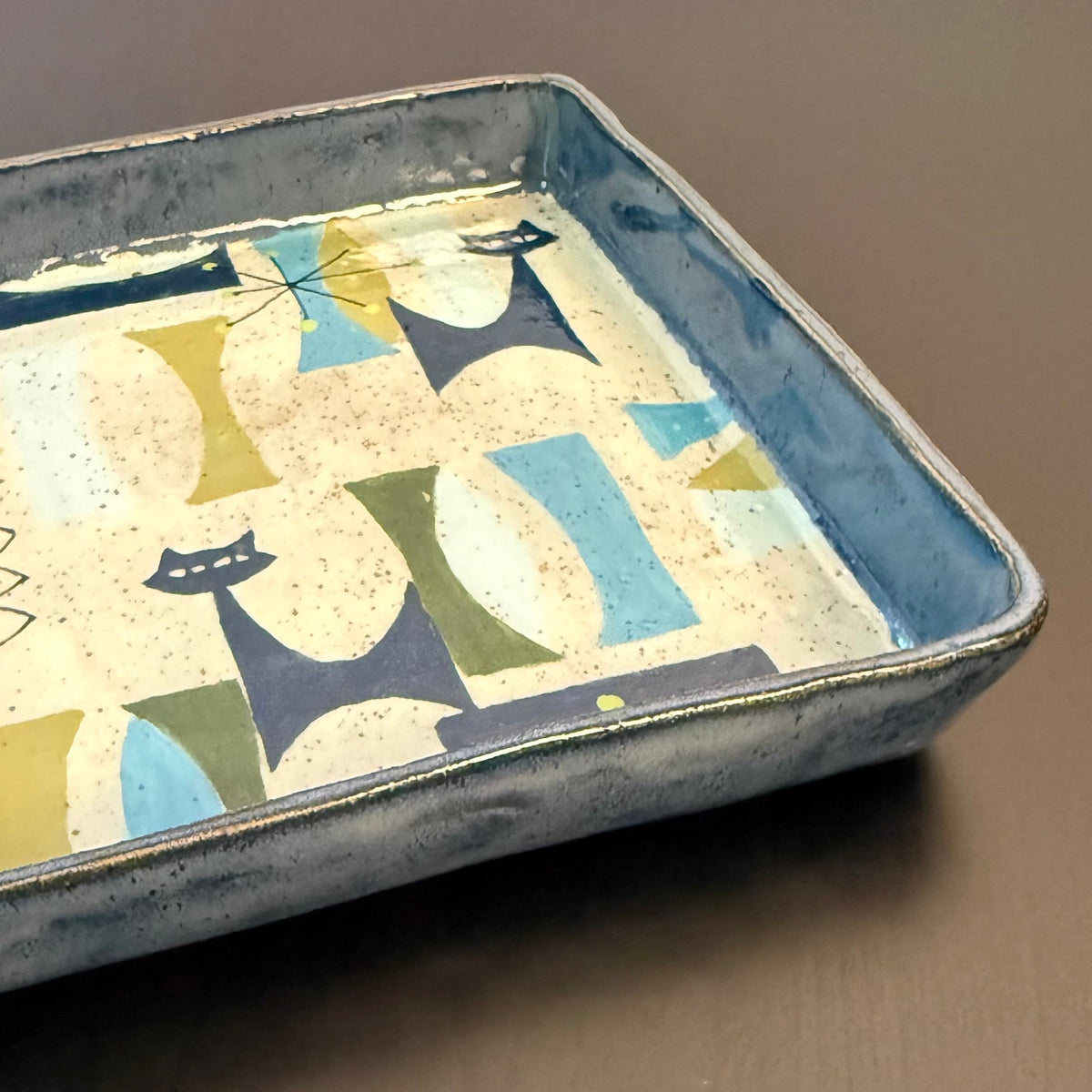 A square ceramic tray with a hand-painted mid-century modern pattern of stylized cats in blue, green, and beige on a cream background, set against a dark wood surface.