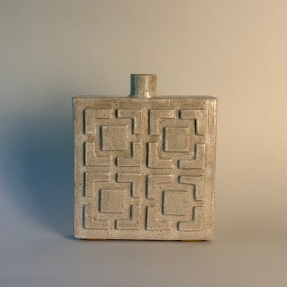 A rectangular ceramic vase with a short cylindrical neck and a raised geometric breeze block pattern on the front, shown on a plain light gray background.