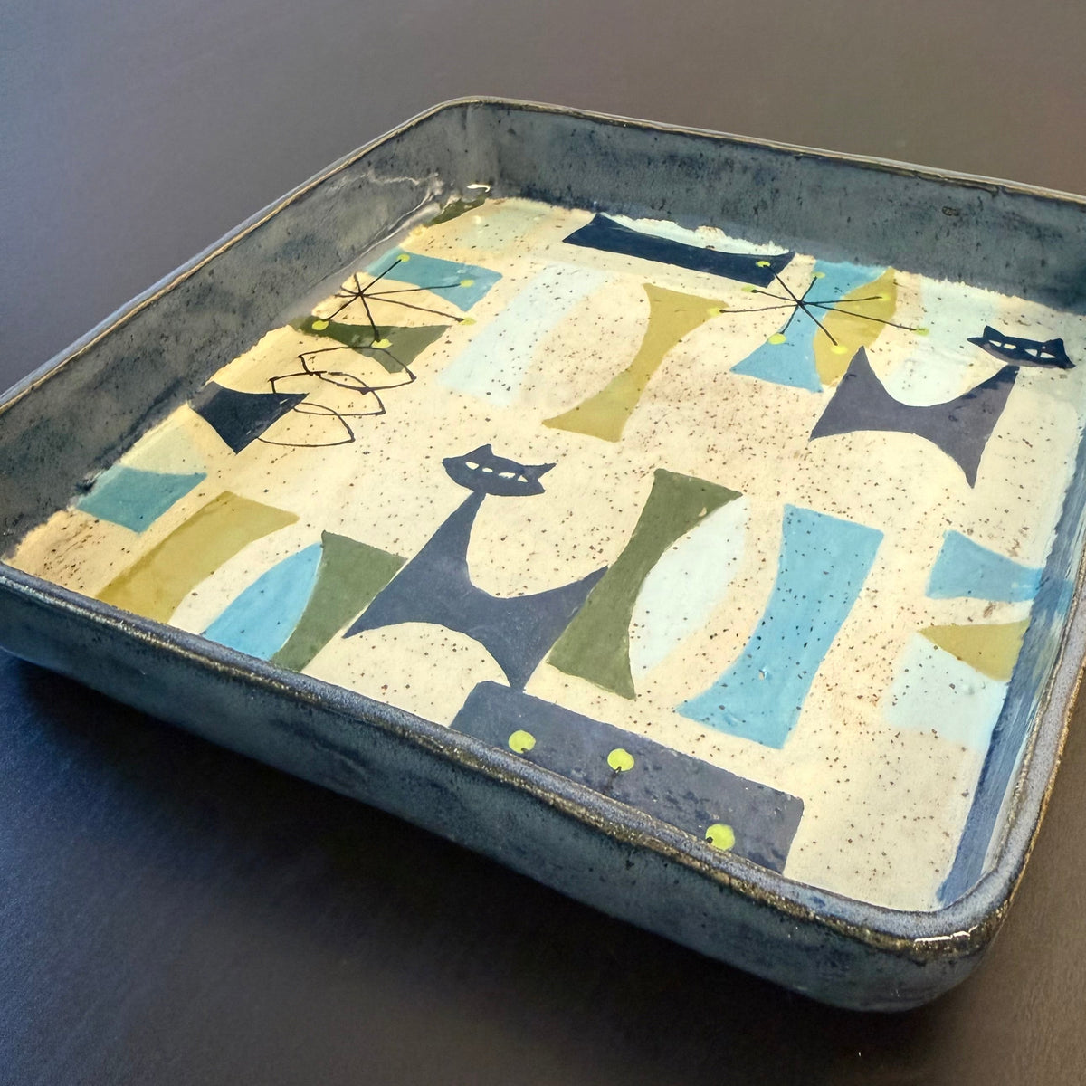 A square ceramic tray with a hand-painted mid-century modern pattern of stylized cats in blue, green, and beige on a cream background, set against a dark wood surface.