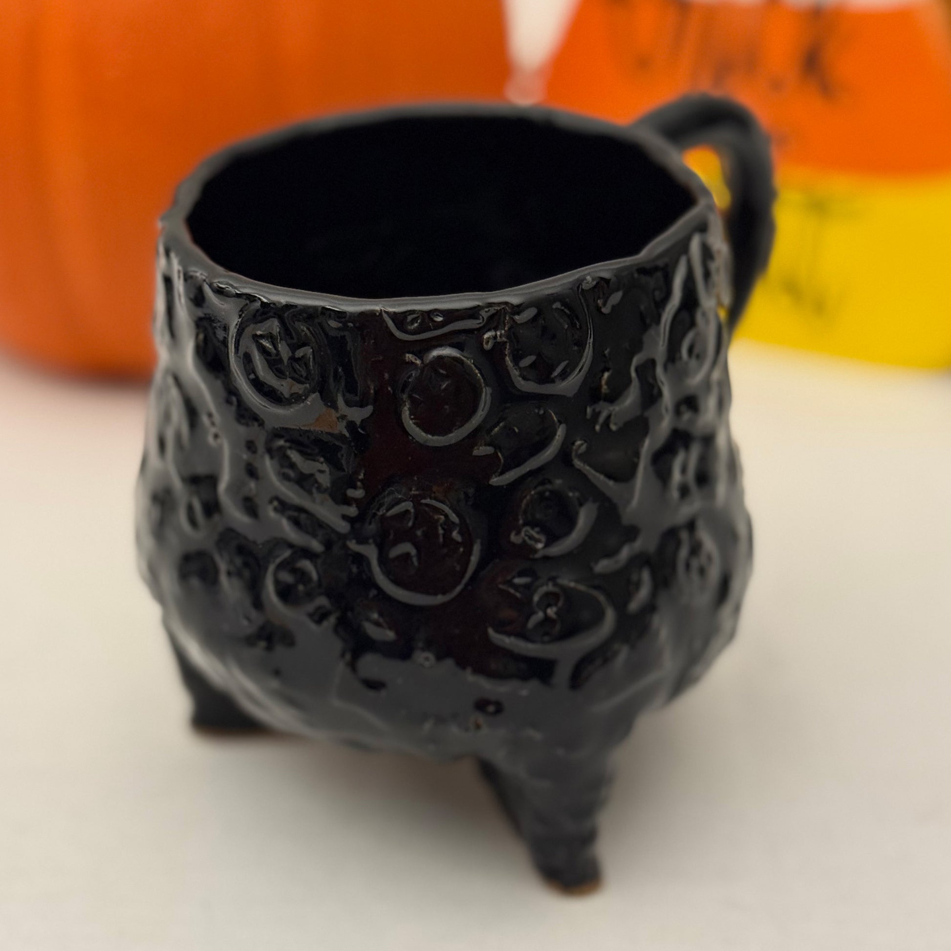 Black Halloween textured cauldron mug on a white surface with a pumpkin in the background.