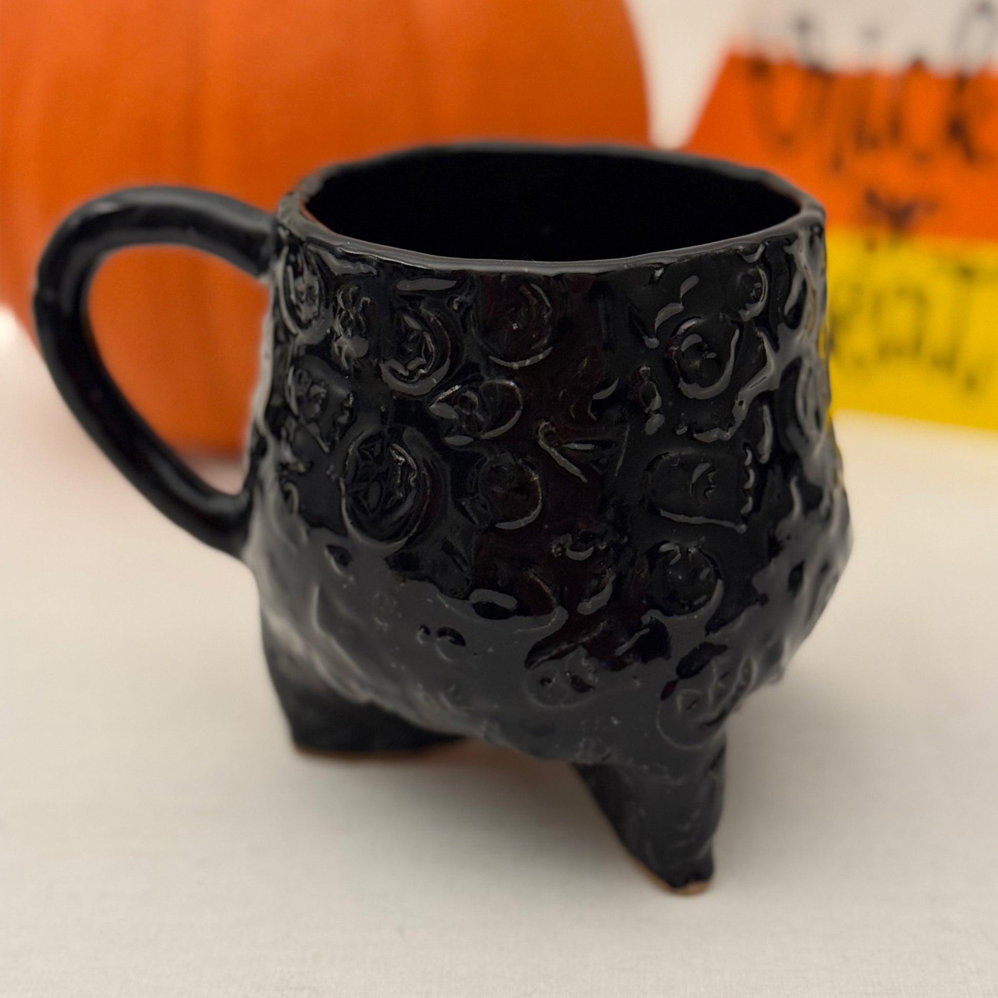 Black Halloween textured cauldron mug on a white surface with a pumpkin in the background.