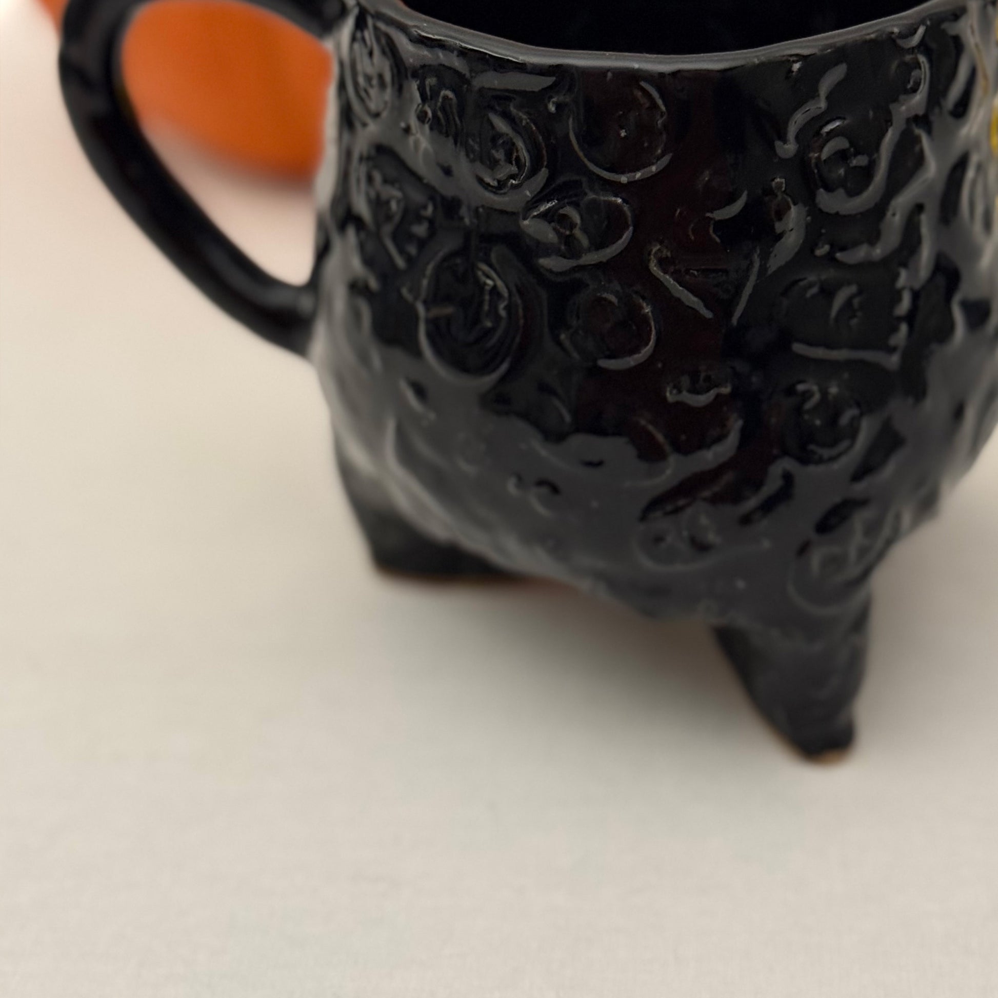 Black Halloween textured cauldron mug on a white surface with a pumpkin in the background.