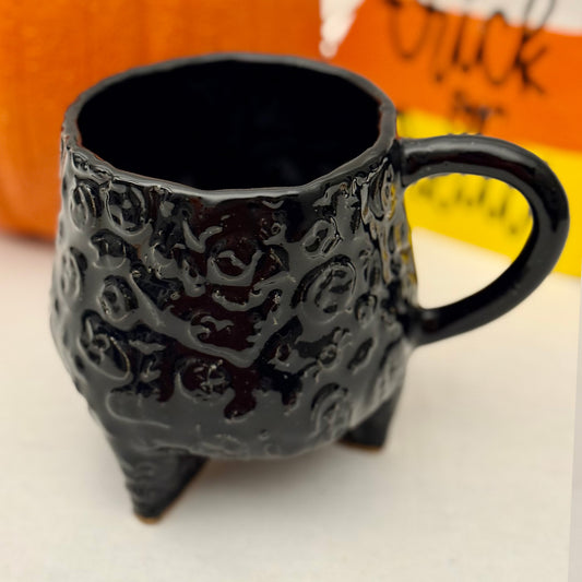 Black Halloween textured cauldron mug on a white surface with a pumpkin in the background.