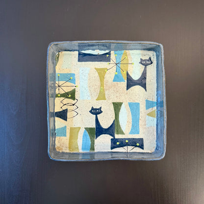 A square ceramic tray with a hand-painted mid-century modern pattern of stylized cats in blue, green, and beige on a cream background, set against a dark wood surface.