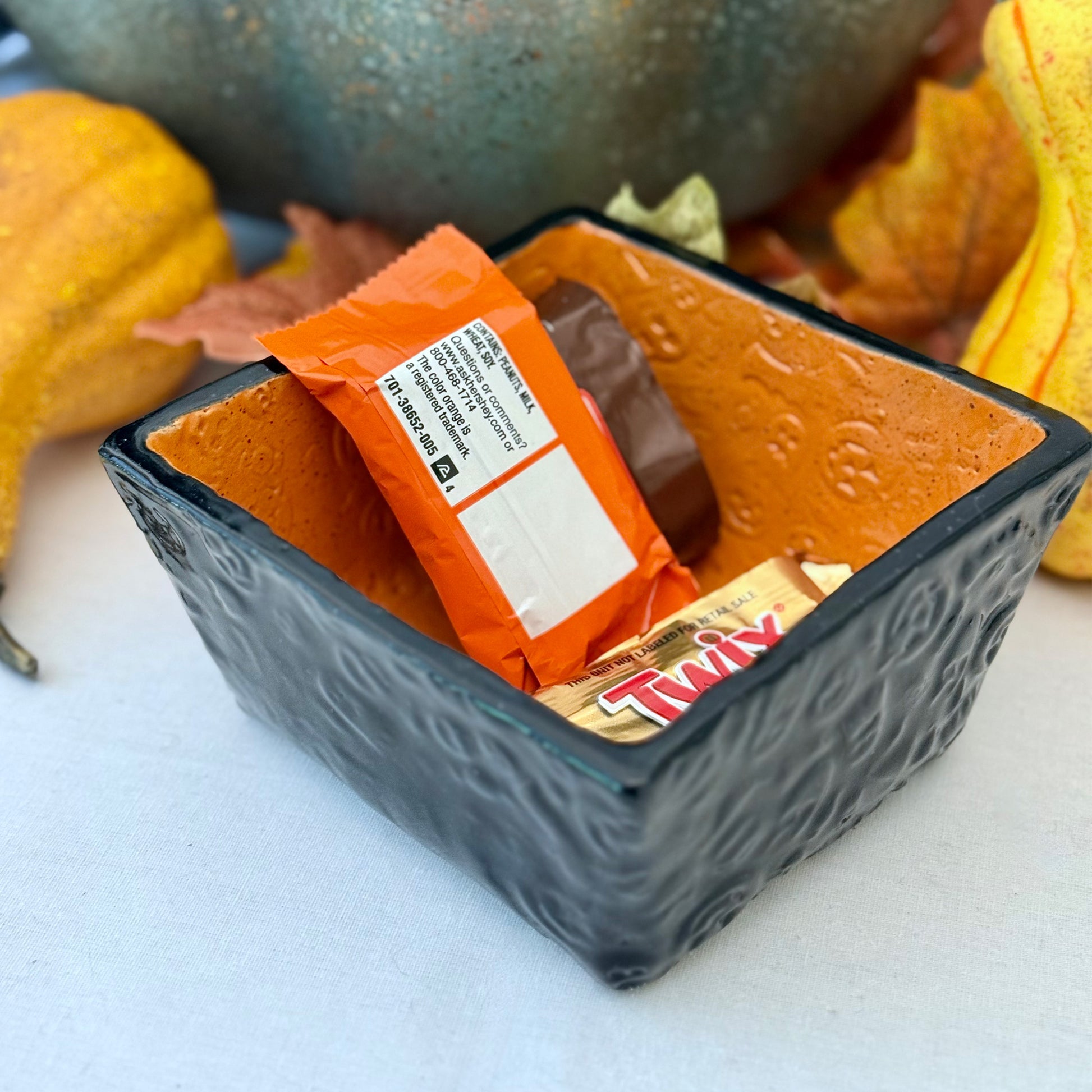Square Halloween textured ceramic candy bowl with candy bars on a white surface with pumpkins in the background.