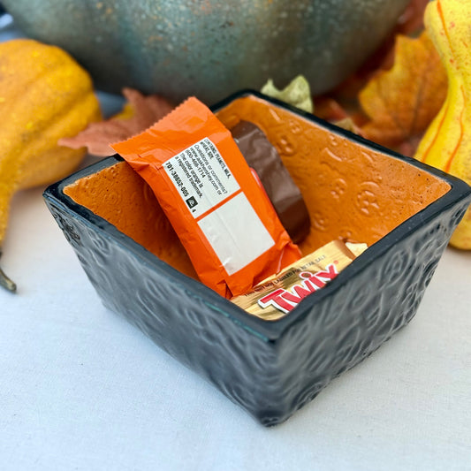 Square Halloween textured ceramic candy bowl with candy bars on a white surface with pumpkins in the background.