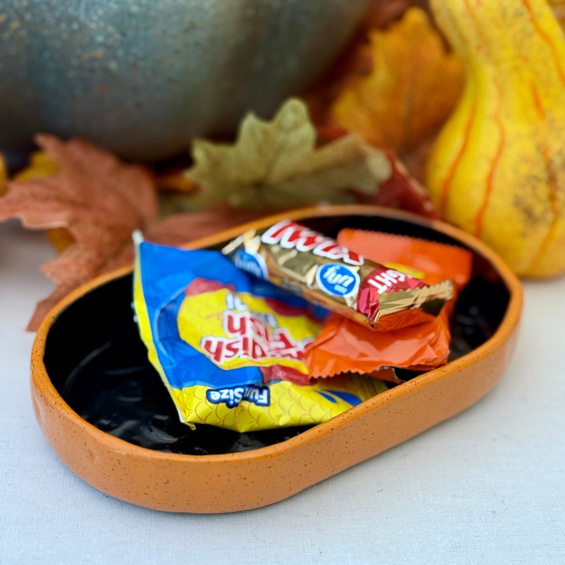 Black and orange Halloween textured oval trinket tray with candy, surrounded by autumn leaves and a small pumpkin.