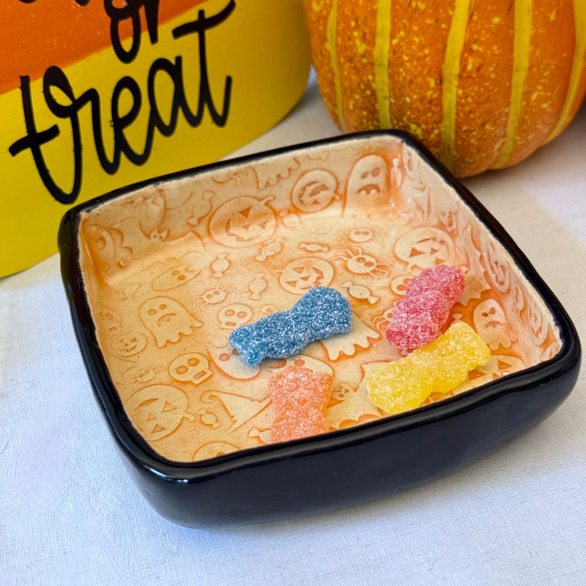 Black bowl with Halloween-themed gummy candies on a white surface with a pumpkin and 'trick or treat' sign in the background.