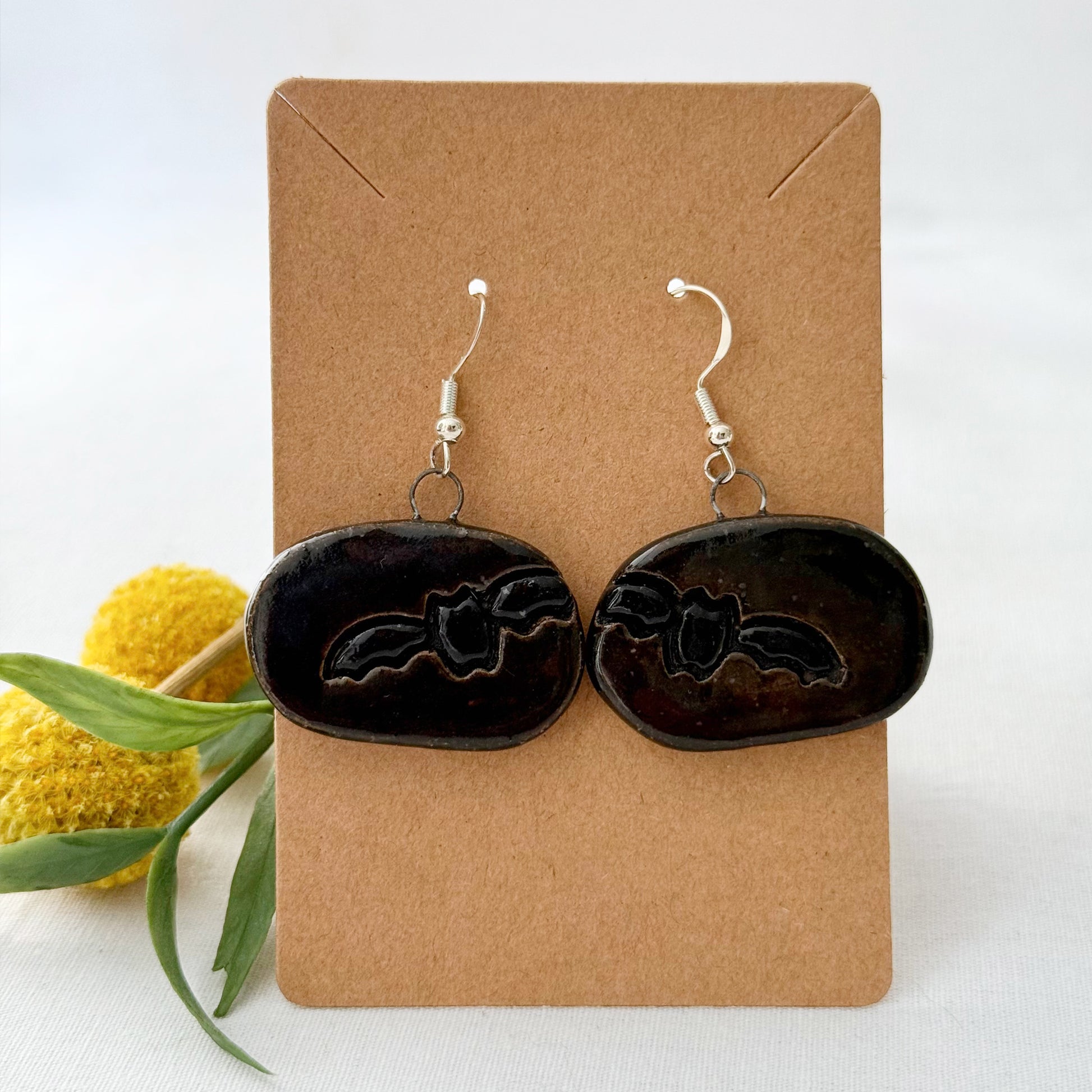 Black bat-shaped earrings on a brown card with yellow flowers and green leaves in the background