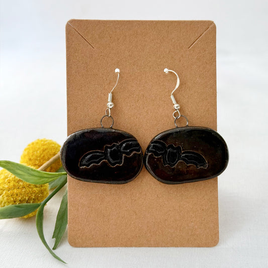 Black bat-shaped earrings on a brown card with yellow flowers and green leaves in the background
