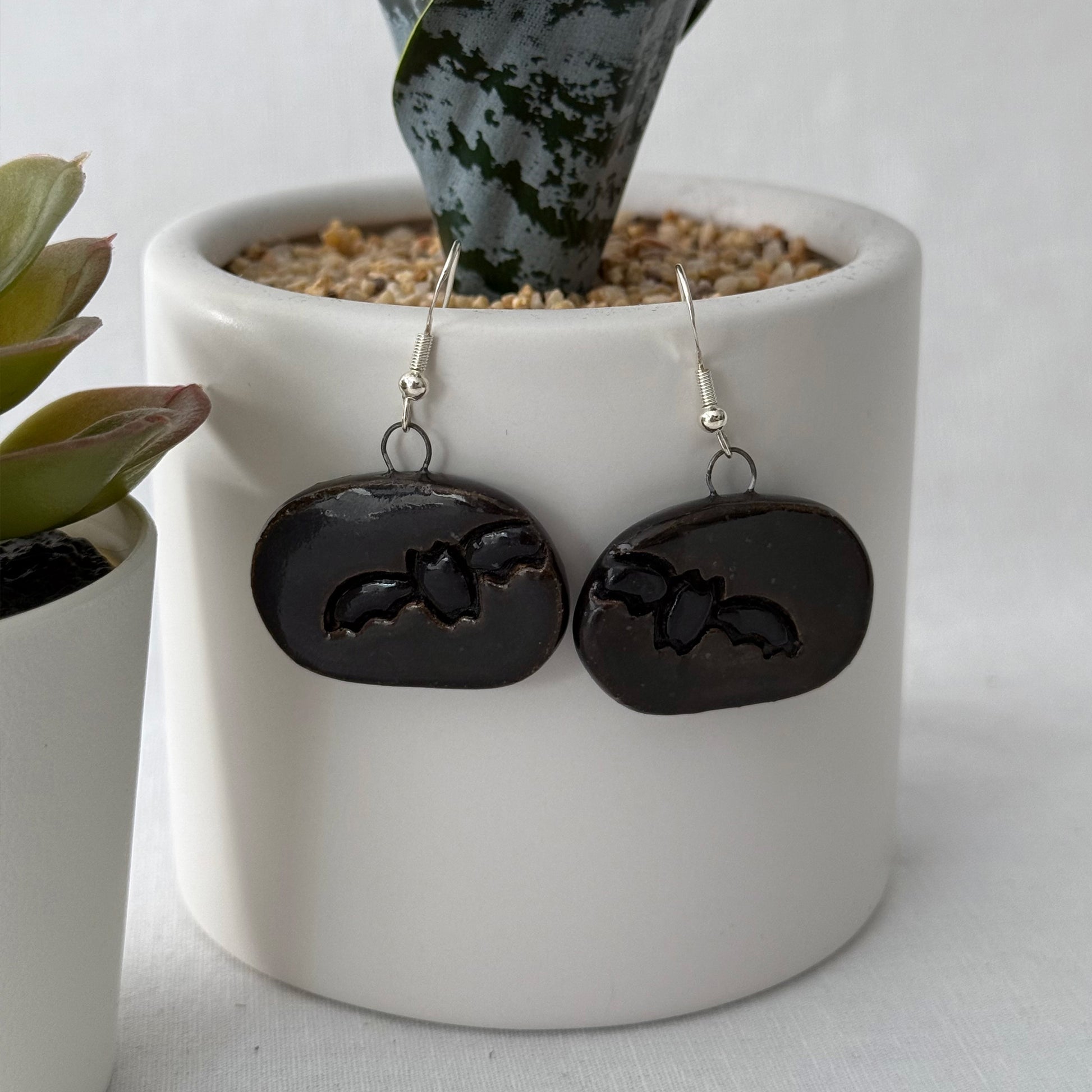 Black bat earrings hanging from a white pot with plants.