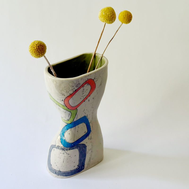 Handmade ceramic vase with interconnected geometric shapes in blue, coral, green, and gray on a sculptural hourglass silhouette.