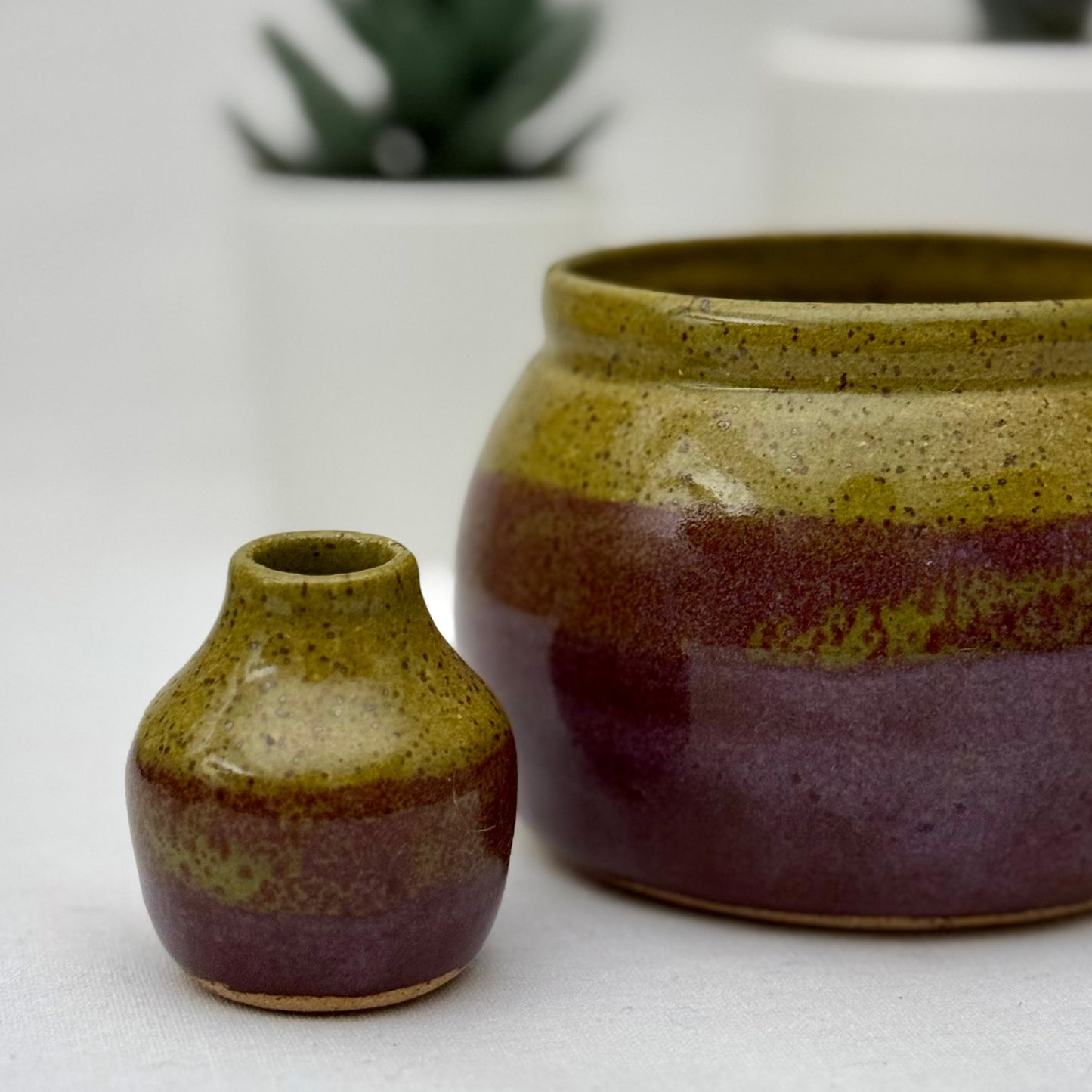 Small olive and purple bud vase in front of a matching pot on a light surface.