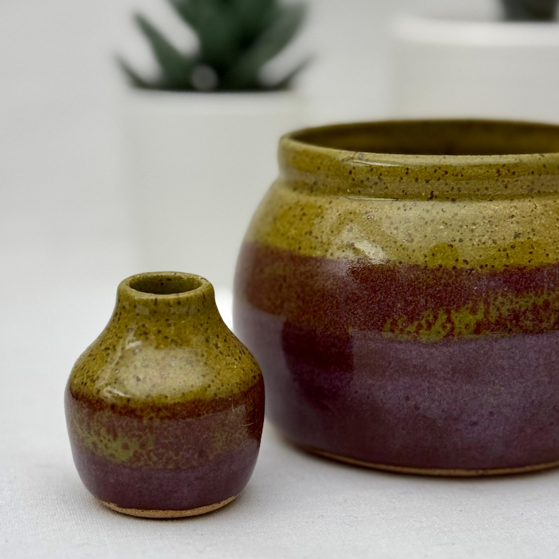 Small olive and purple bud vase in front of a matching pot on a light surface.