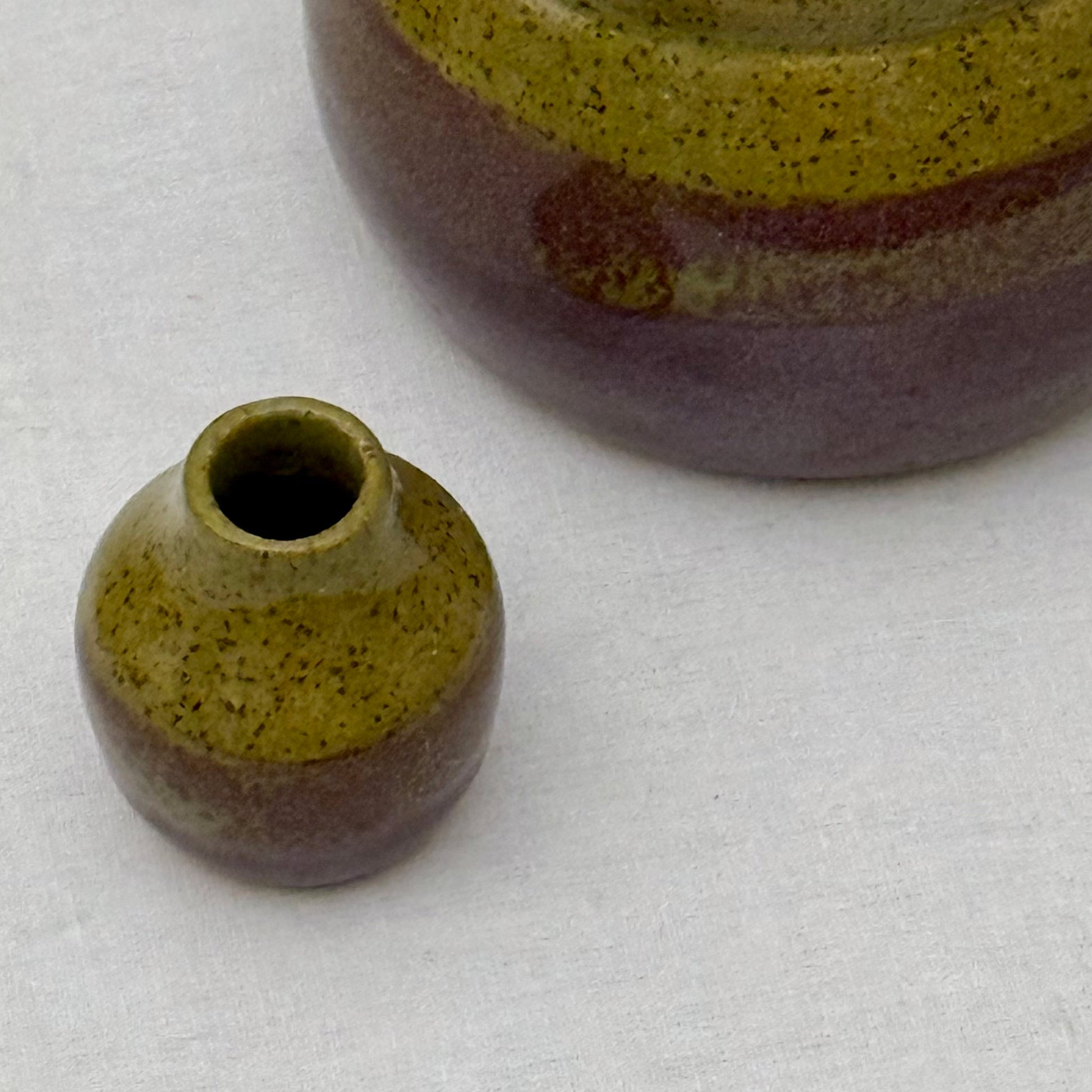 Small olive and purple bud vase in front of a matching pot on a light surface.