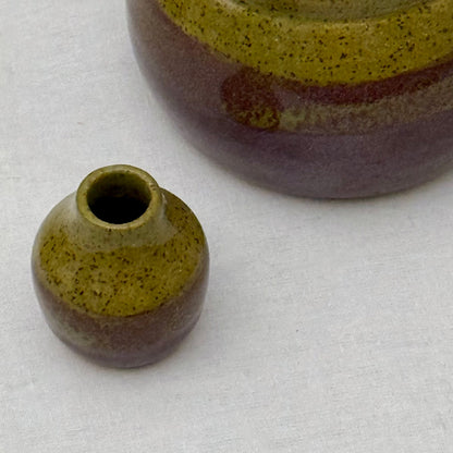 Small olive and purple bud vase in front of a matching pot on a light surface.
