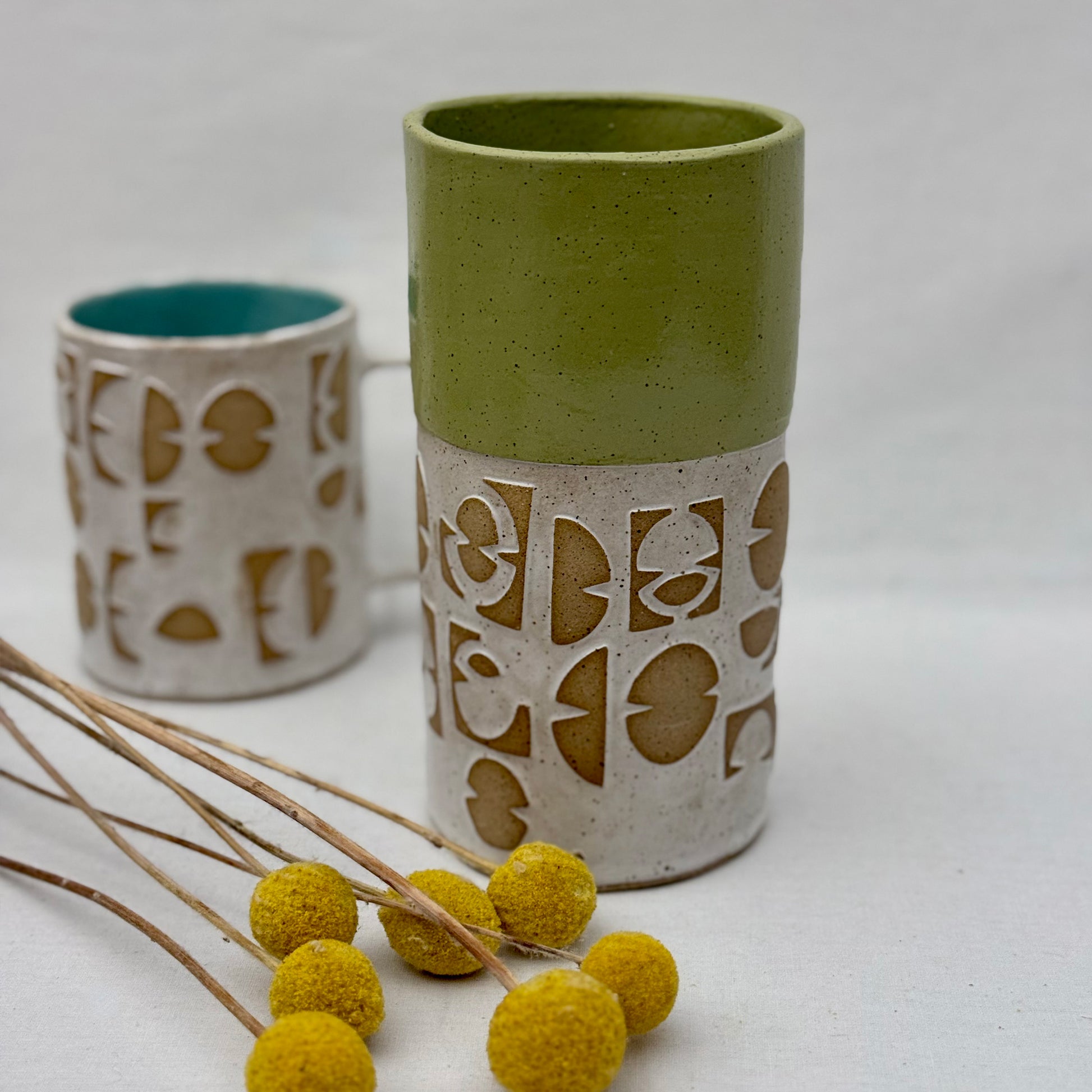 ceramic vase with geometric patterns with matching mug behind it and a small arrangement of yellow flowers on a white background