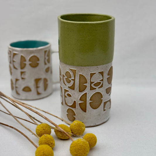 ceramic vase with geometric patterns with matching mug behind it and a small arrangement of yellow flowers on a white background