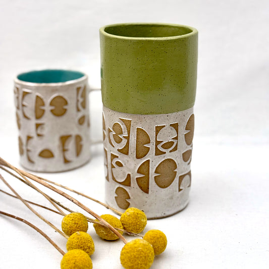 Ceramic vase with mid century inspired patterns with matching mug behind it and a small arrangement of yellow flowers on a white background